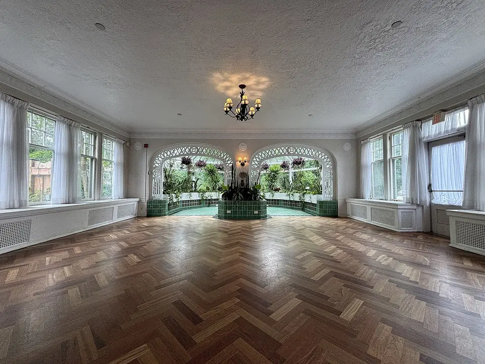Véranda intérieure lumineuse bordée de fenêtres à petits bois, avec parquet à chevrons et arcades vitrées ouvrant sur un jardin d'hiver.